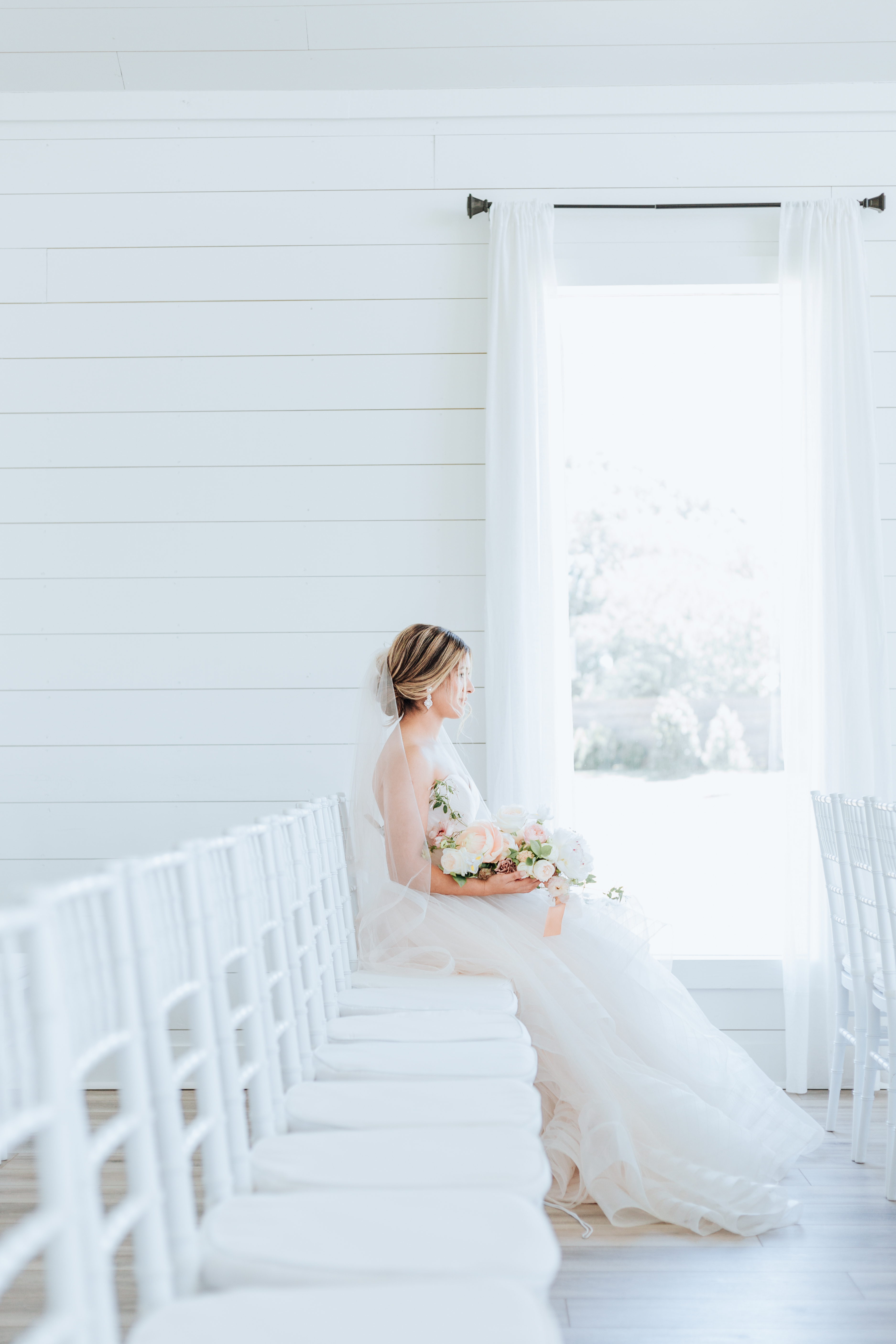 a woman in a wedding dress seated by a window with a wedding bouquet of flowers photographed by joanna booth weddings