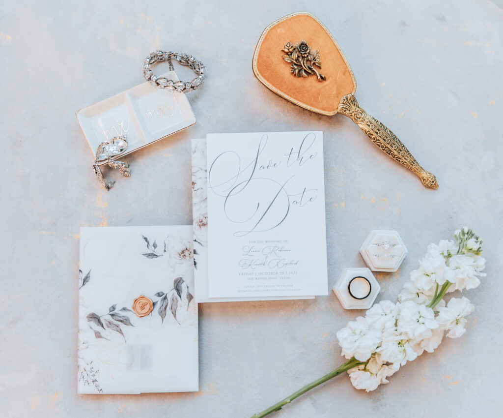 Joanna Booth Weddings captures the flat lay details at the Peach Orchard Venue in The Woodlands. The flatlay includes a brush, wedding rings, save the date cards, and wedding jewelry